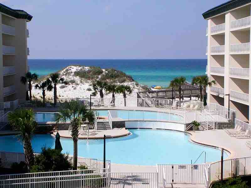 A view of the pool area surrounded by palm trees, with a private beach access in the background. The calm blue waters of the Gulf can be seen beyond the pool, framed by sandy dunes and a clear sky.