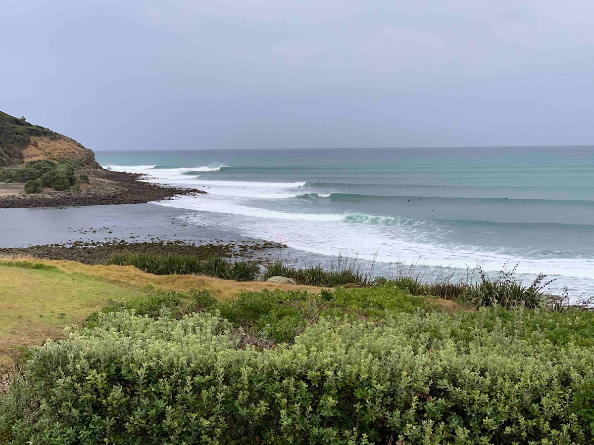 A view of the coastline showcases rolling waves breaking gently on the shore. Lush greenery borders the foreground, while a rocky outcrop extends towards the water. The ocean's varying shades of blue are visible under a cloudy sky.