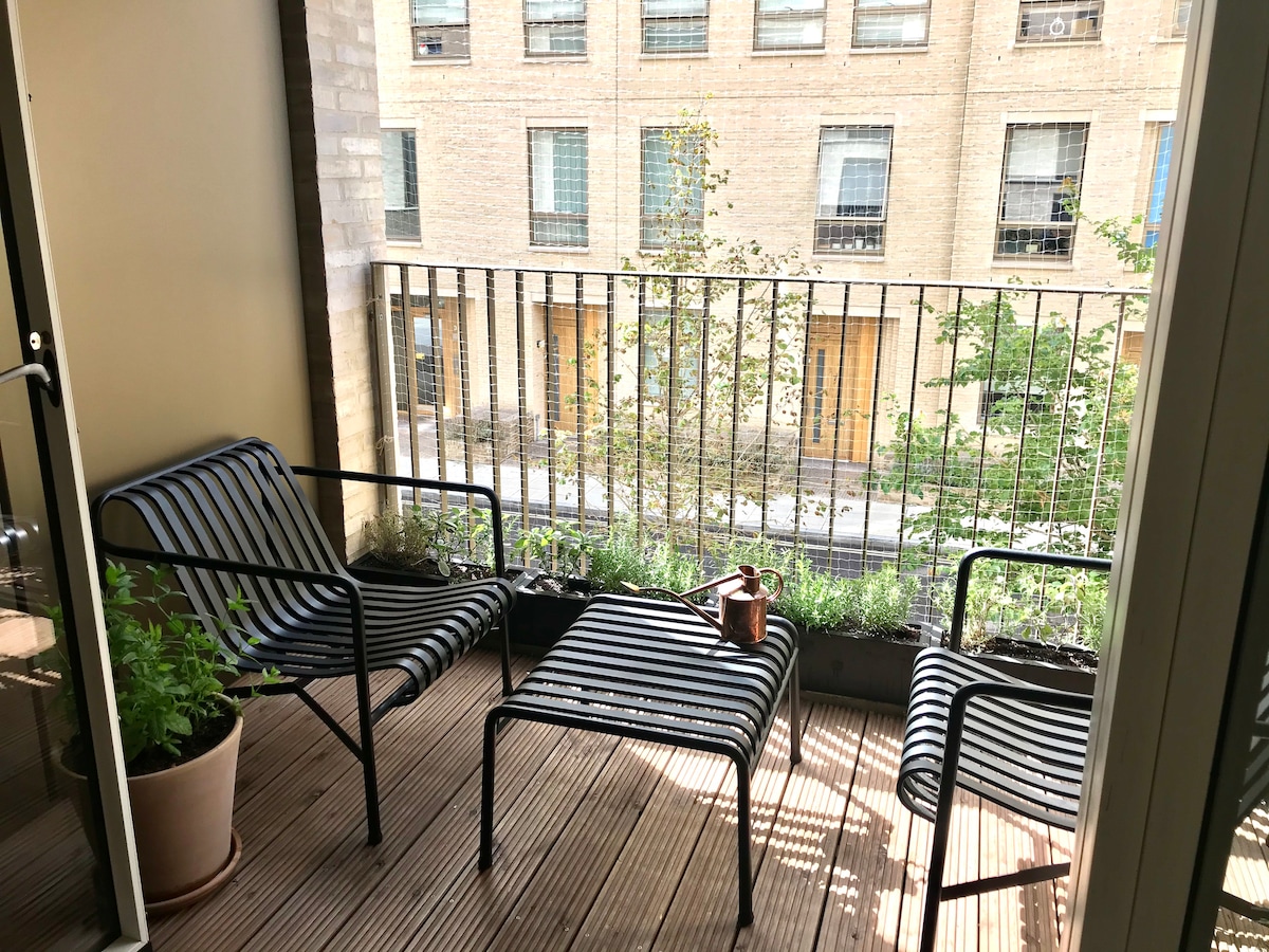 A private balcony features modern outdoor furniture, including two lounge chairs and a coffee table. Green plants are positioned in planters along the railing, providing a refreshing touch. The space offers a view of the surrounding greenery and building's exterior.