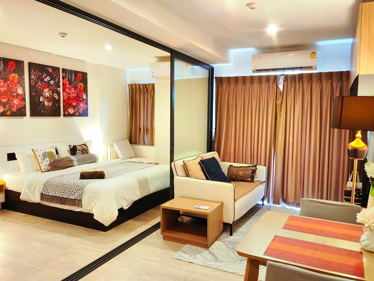 An airy room features a spacious bed and a sofa bed, arranged thoughtfully within the layout. Floor-to-ceiling glass mirrors add depth, while soft fabrics dress the windows and a dining area is provided. Artwork depicting red flowers adds a splash of color to the neutral decor.