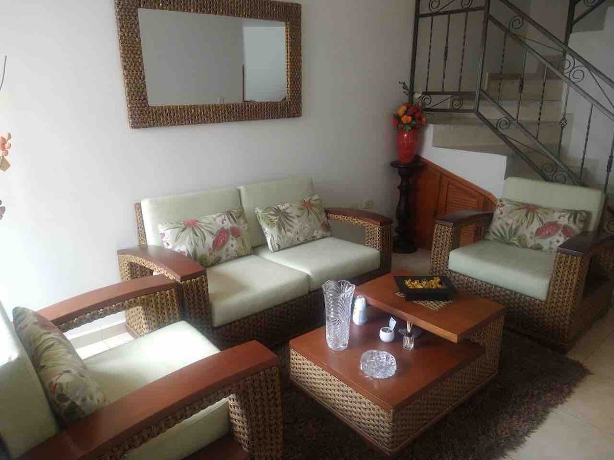 A bright and airy living area features a set of comfortable seating with light-colored cushions and woven detailing. A wooden coffee table occupies the center space, adorned with decorative items. Natural light enters through a nearby window, reflecting off a large mirror on the wall.