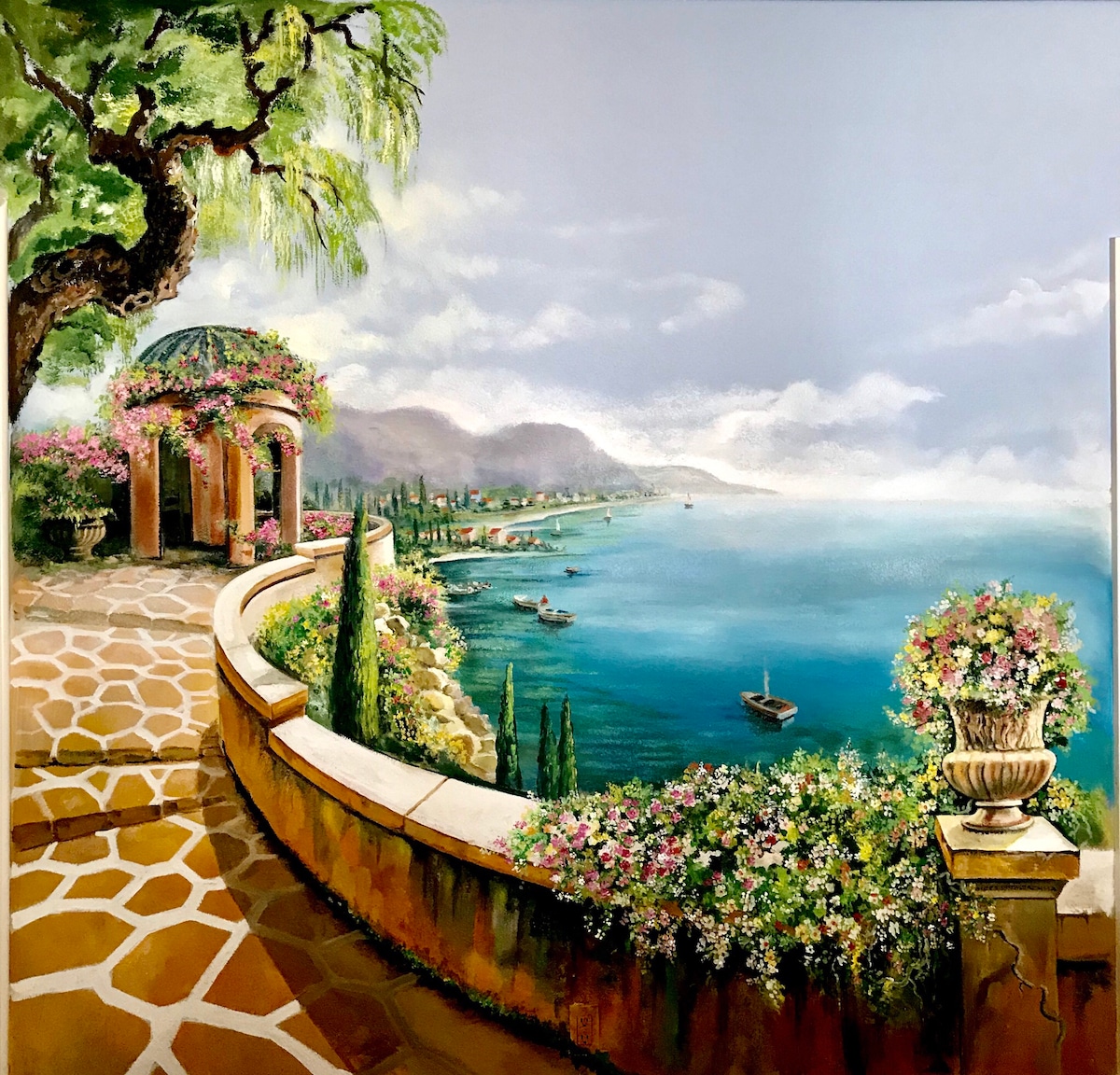 A scenic view features a winding pathway bordered by blooming flowers, leading to a coastline with distant boats. The horizon showcases gentle hills and a tranquil sea under a softly illuminated sky, while stone walls surround the lush greenery, creating a serene atmosphere.