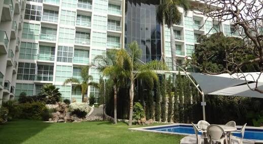 A multi-story building with extensive glass windows is surrounded by manicured greenery. A small pool and seating area are visible in the foreground, complemented by palm trees and a shaded area, reflecting a serene outdoor space ideal for relaxation.