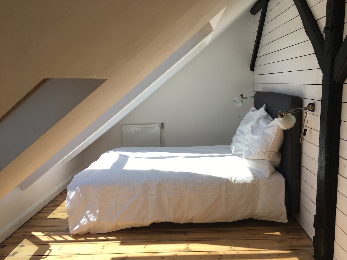 A light-filled sleeping area features a spacious bed adorned with fresh white linens. A gentle slope of the ceiling is accented by exposed wooden beams. Wall-mounted lamps provide soft lighting, while a radiator is positioned conveniently nearby.