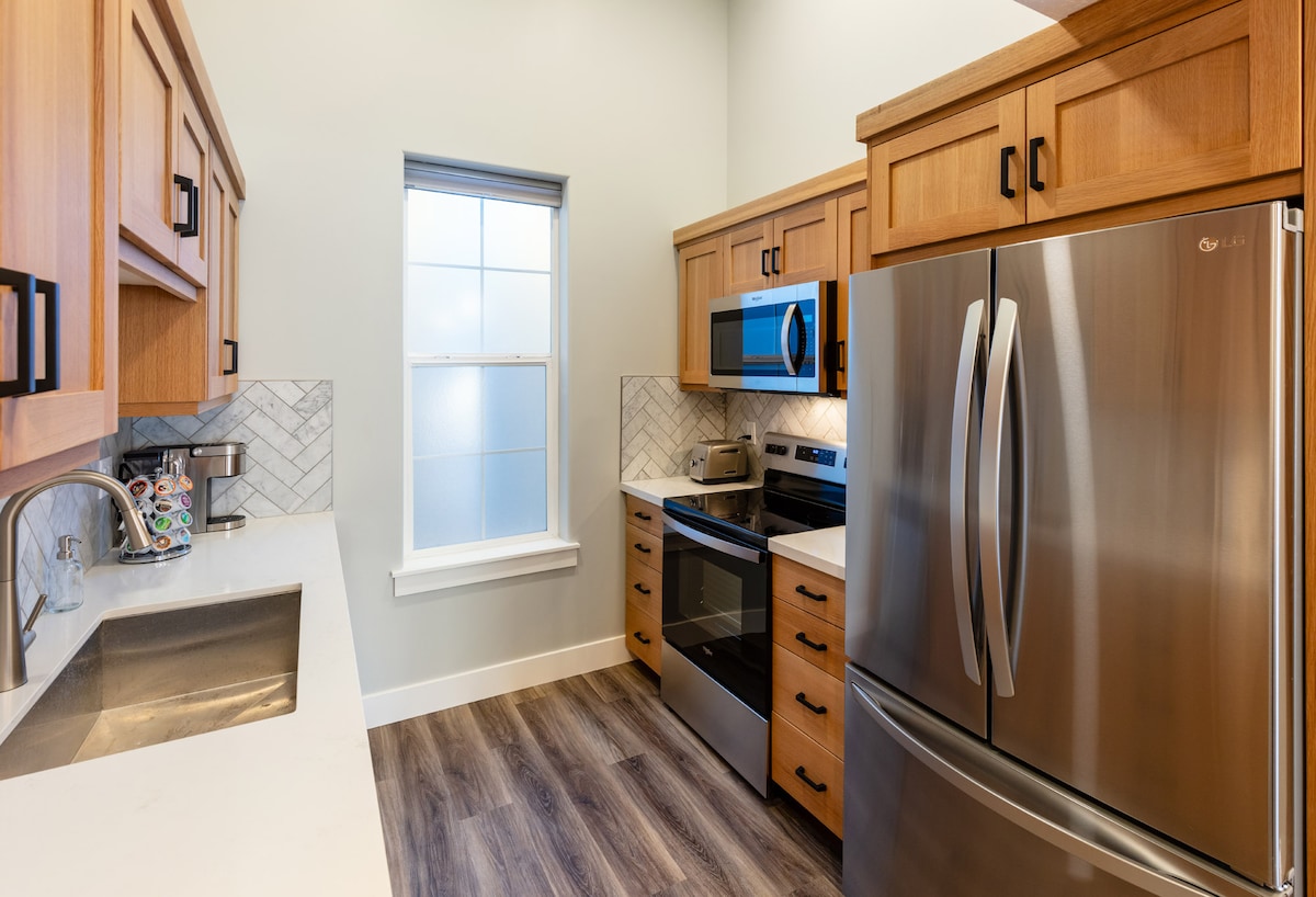 A full kitchen features wooden cabinets and stainless steel appliances, including an oven, microwave, and refrigerator. A sink with a modern faucet is positioned beneath a window, allowing natural light to illuminate the space. The floor is adorned with wood-look tiles.