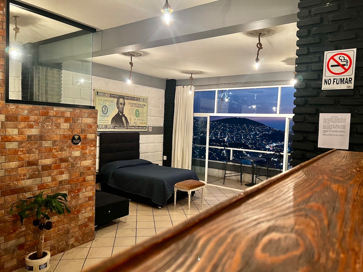 A modern room features a dark-colored bed with a matching blanket, positioned near large windows offering panoramic views of a cityscape at night. Brick and glass walls enhance the contemporary design, while a small table and plant add subtle touches of comfort.
