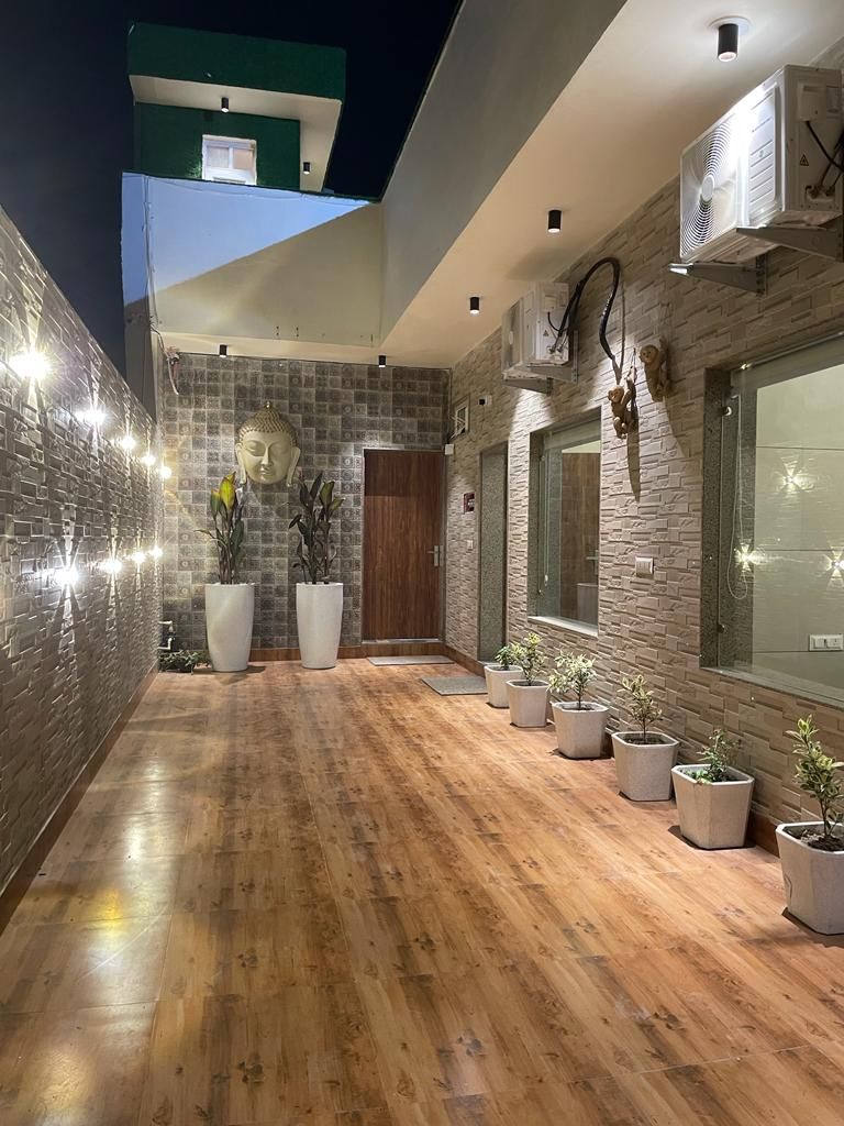 A well-lit corridor features modern stone walls and a polished wooden floor. Large potted plants line the sides, adding greenery to the space. Wall-mounted lighting enhances the ambiance, while the main entrance is visible at the far end, offering a welcoming entry point.