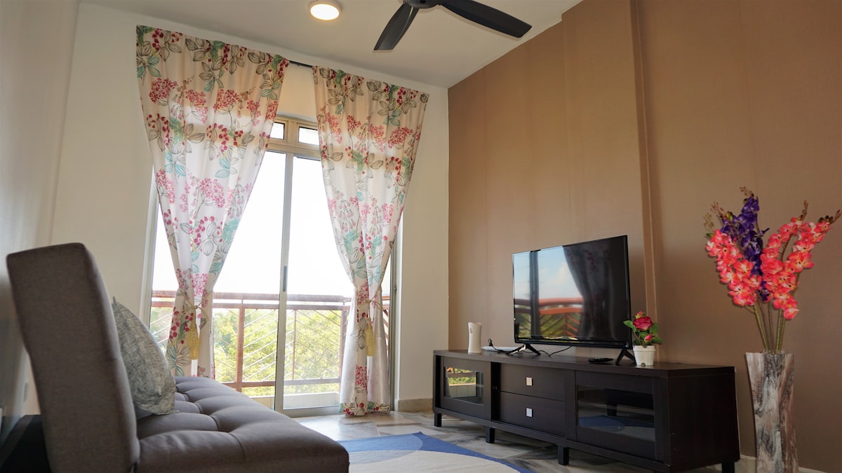 A cozy living area is presented, featuring large windows adorned with floral curtains that allow natural light to fill the space. A comfortable sofa sits opposite a flat-screen television, complemented by a decorative table with flowers. A ceiling fan is mounted above for added comfort.