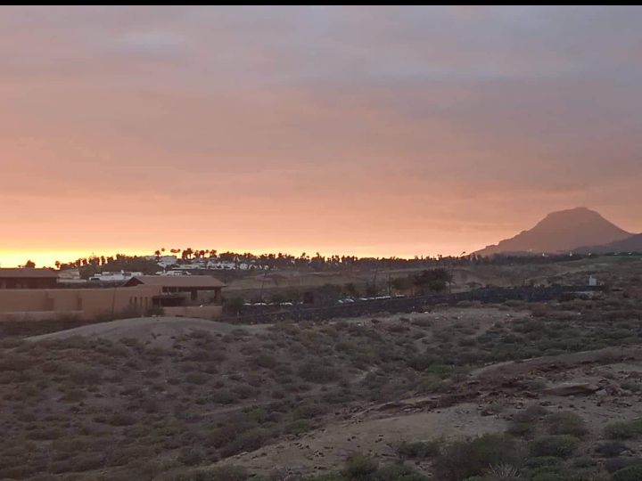 A scenic view of the landscape at sunset shows soft pastel hues in the sky, complemented by a distant mountain outline. The foreground features natural terrain with sparse vegetation, creating a serene backdrop against the warm colors of the setting sun.