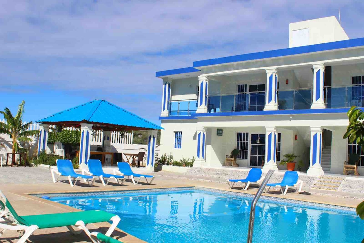 A two-story building is framed by lush greenery, featuring blue accents along its columns and roof. A swimming pool is situated in the foreground, surrounded by lounge chairs. An outdoor seating area under a blue-roofed gazebo can be seen to the left.