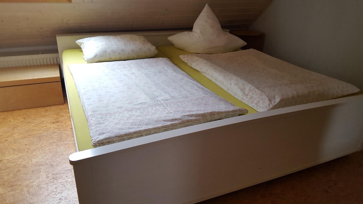 A spacious bed is positioned in a softly lit room, featuring light-colored bedding with floral patterns. Two pillows rest at the head, and one large duvet is neatly spread across the mattress. Minimalistic wooden furniture lines the walls, enhancing the serene atmosphere.