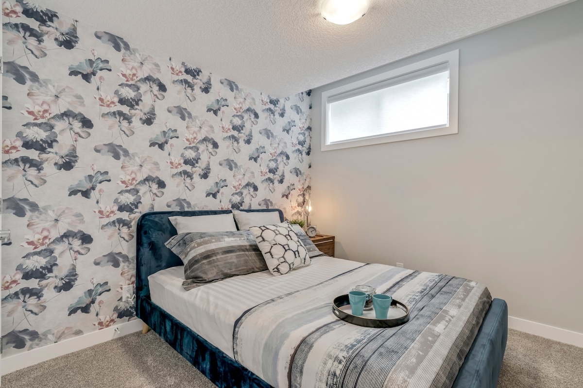 A cozy bedroom showcases a spacious bed layered with patterned bedding and decorative pillows. The serene floral wallpaper complements the soft grey tones, while a small nightstand holds two glasses and a plate. A large window provides natural light, enhancing the calming atmosphere.