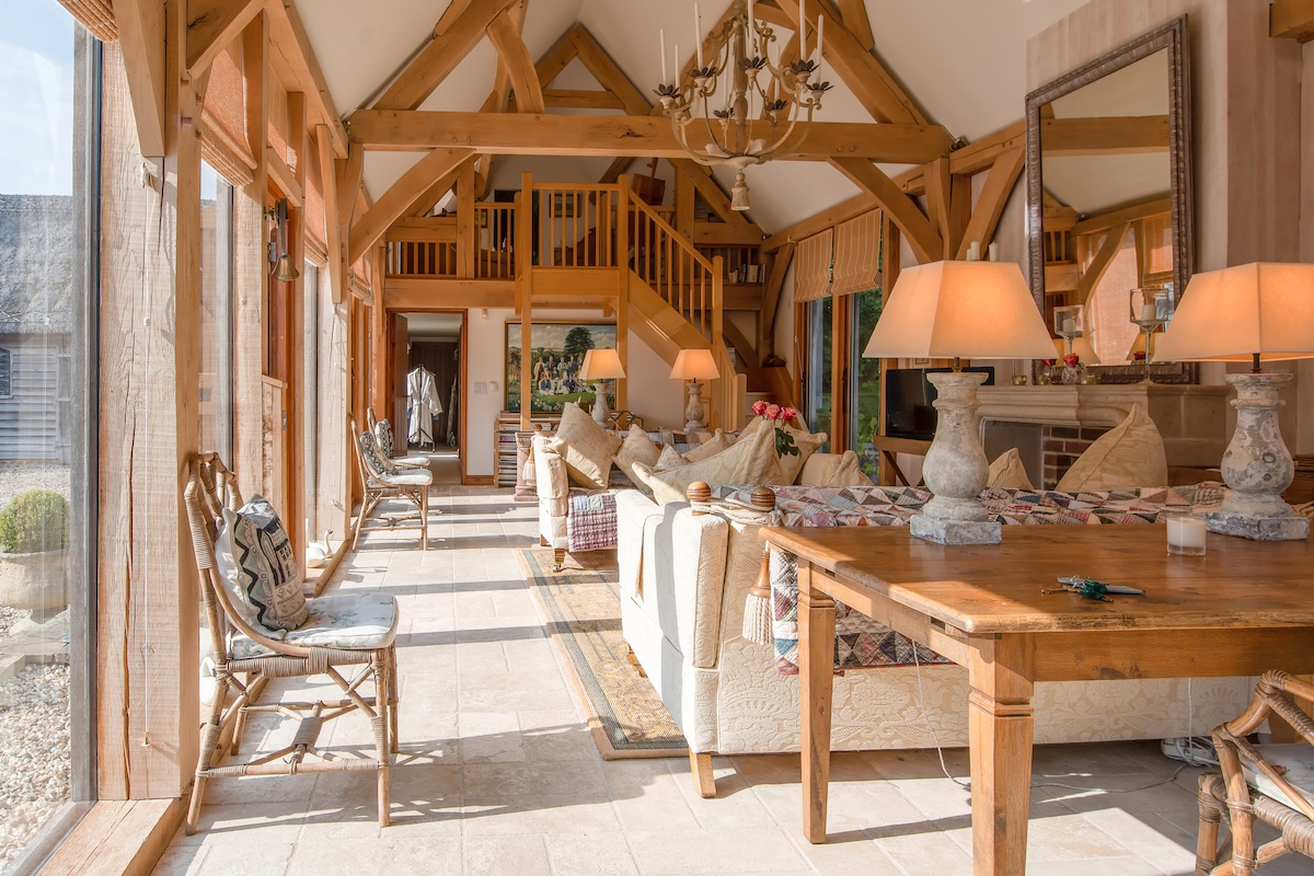 A spacious living area is highlighted by inviting oak beams and large windows that allow natural light to fill the space. Comfortable seating arrangements with soft cushions are complemented by a central dining area, enhancing the open layout. Elegant lighting fixtures add to the ambiance.