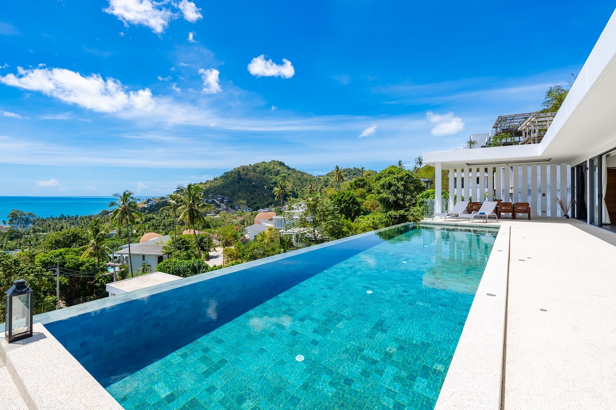 An expansive infinity pool overlooks lush greenery and distant hills leading to the ocean. Sunbeds are positioned beside the pool, inviting relaxation under clear blue skies. The modern villa structure features large glass doors that connect the living area with the outdoor space.