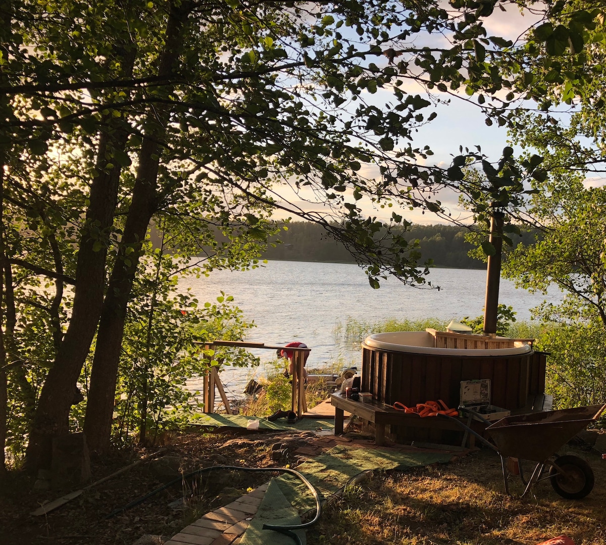 A wooden hot tub is positioned among lush greenery, with a serene lake visible in the background. Sunlight filters through the trees, casting gentle shadows on the ground. A pathway leads toward the water, enhancing the peaceful natural setting.