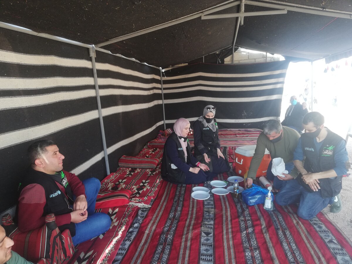 A spacious tent is decorated with traditional black and white striped fabric. Several people sit on woven carpets, engaging in conversation. Plates and utensils are arranged on a low table. Natural light enters through the open entrance, illuminating the inviting atmosphere.