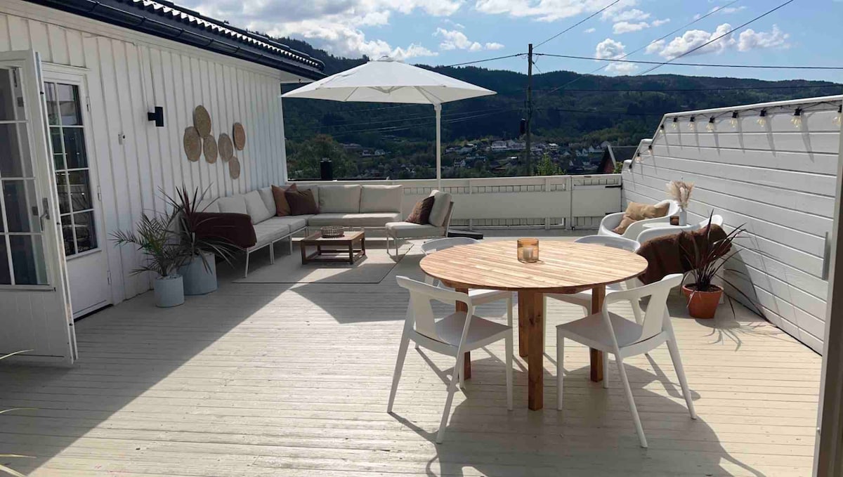 An expansive terrace is featured, equipped with a round wooden table surrounded by four white chairs. A spacious seating area with light-colored cushions is positioned next to a large umbrella, offering shade. Scenic views are visible in the background.