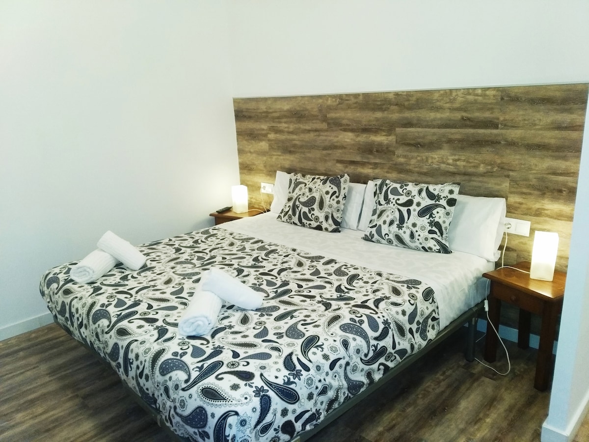 A spacious bed is presented with a patterned duvet and complementary pillows. Nightstands with soft lighting flanking the bed create a balanced aesthetic. Fresh towels are neatly placed at the foot of the bed, contributing to the inviting presentation.