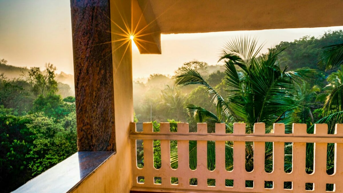 A balcony view showcases lush greenery with palm trees set against a golden sunrise. The soft morning light filters through the treetops, creating a serene atmosphere. The balcony's railing features a simple design, complementing the natural scenery.
