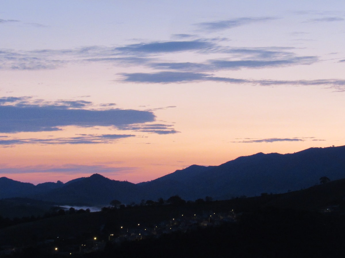 A serene view of mountains during dawn, featuring soft pastel colors in the sky. The silhouette of the hills contrasts with the gentle light emerging from the horizon, while distant lights from the village below add a hint of warmth to the scene.