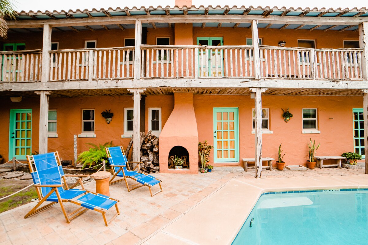 A courtyard area features an inviting swimming pool surrounded by terracotta tiles. Two blue lounge chairs are positioned nearby, with a fireplace built into the wall. The exterior walls of the building are painted in a warm orange hue, complemented by turquoise door frames.
