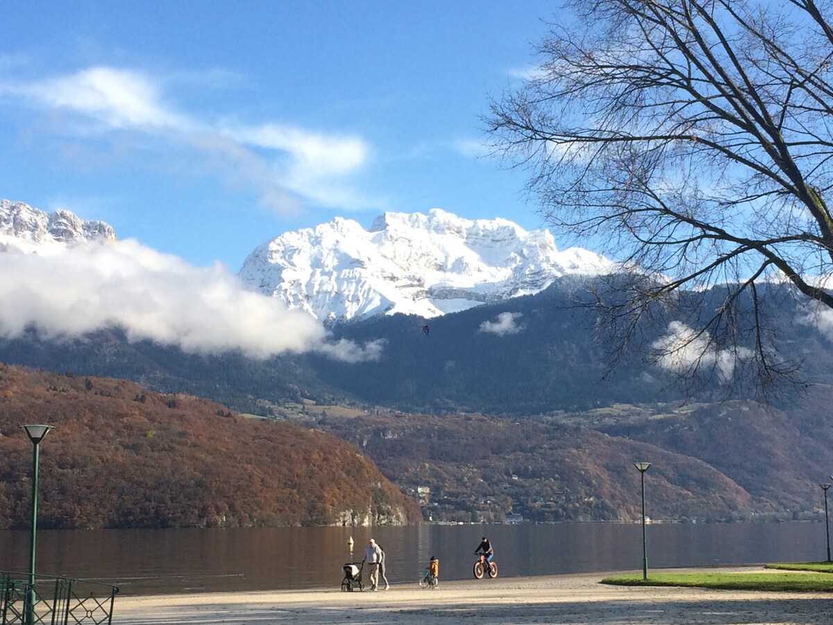 A scenic view of a serene lake surrounded by mountains with snow-capped peaks. A pathway along the water's edge is visible, where individuals can be seen biking and walking. The landscape reflects a calm atmosphere under a clear blue sky with wisps of clouds.