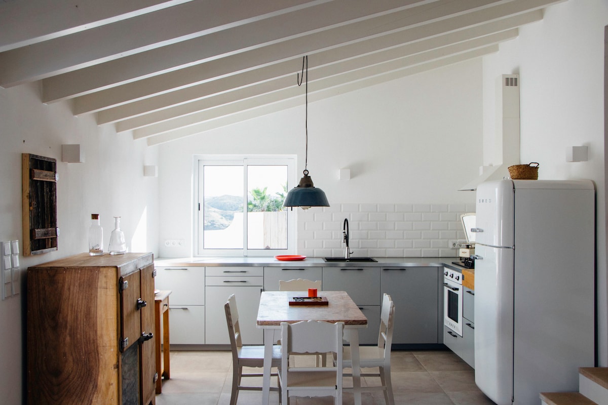A modern kitchen scene features high, slanted ceilings with natural light streaming through a large window. The space includes a wooden dining table surrounded by white chairs, a refrigerator, and an equipped cooking area with a stove and sink. Neutral tones create a clean and airy atmosphere.