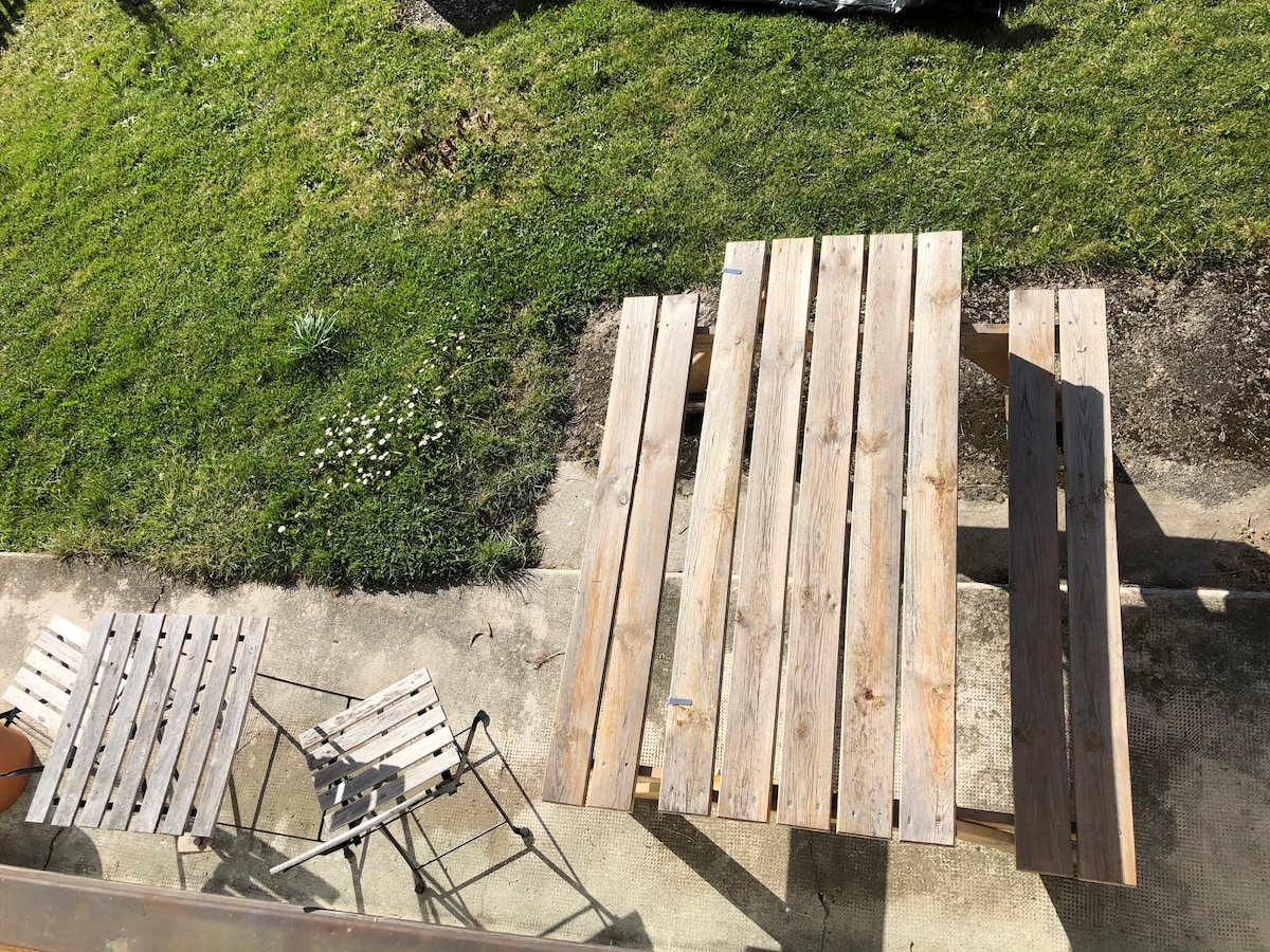 An outdoor area features a weathered wooden table accompanied by two folding chairs. The scene is surrounded by green grass and a few scattered flowers, providing a serene setting for relaxation. The layout suggests a peaceful space for enjoying meals outside.