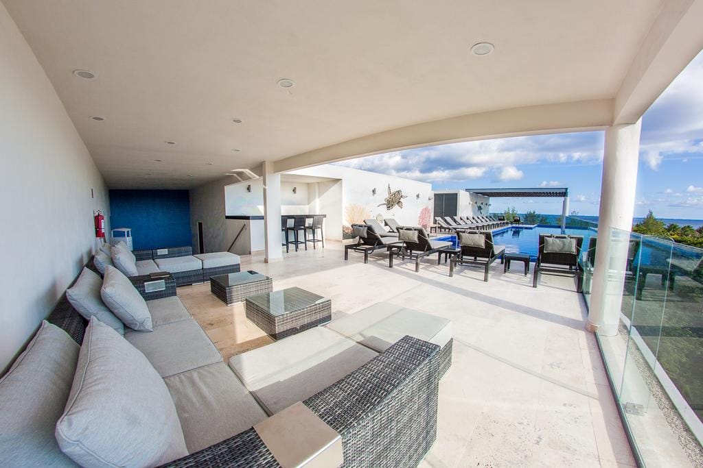 A spacious outdoor terrace is shown, furnished with multiple seating options including a large sectional sofa and lounge chairs. A dining area is visible in the background, and a pool is positioned beyond the glass railing, offering expansive views of the surroundings.