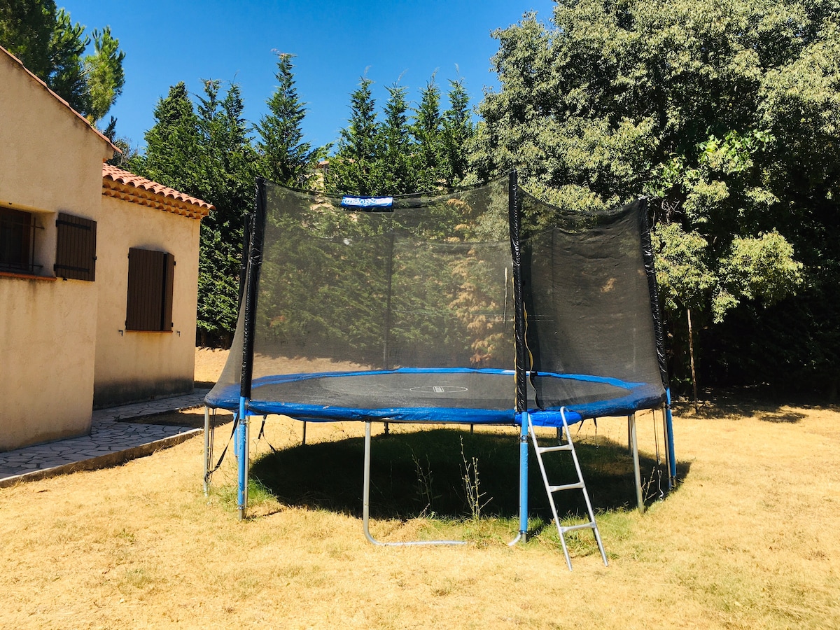 A spacious trampoline is set in a grassy area surrounded by trees, providing a safe space for children to play. The trampoline features a protective net and a ladder for easy access. The adjacent house, with brown window shutters, is partially visible.