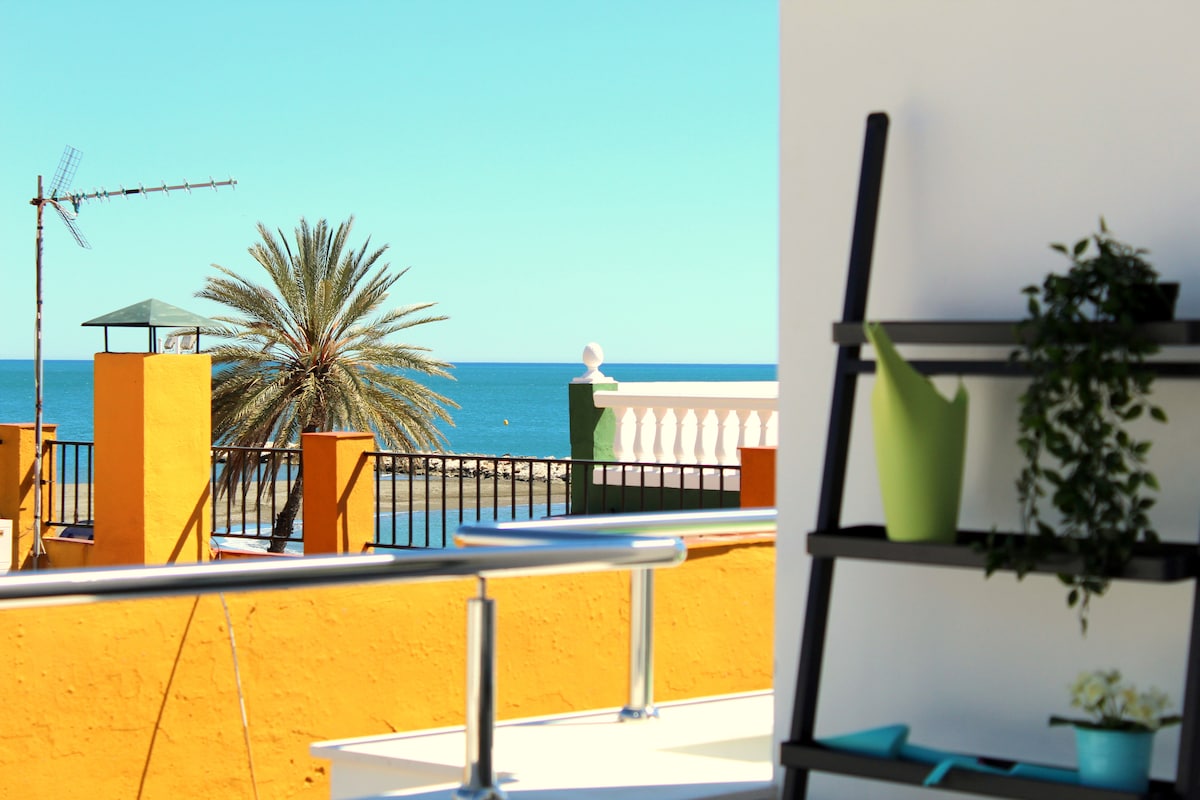 A view is presented from a balcony overlooking the sea, framed by a vibrant orange wall. A palm tree is visible in the background, with a clear blue sky and serene waters. A modern shelf with greenery is positioned in the foreground.