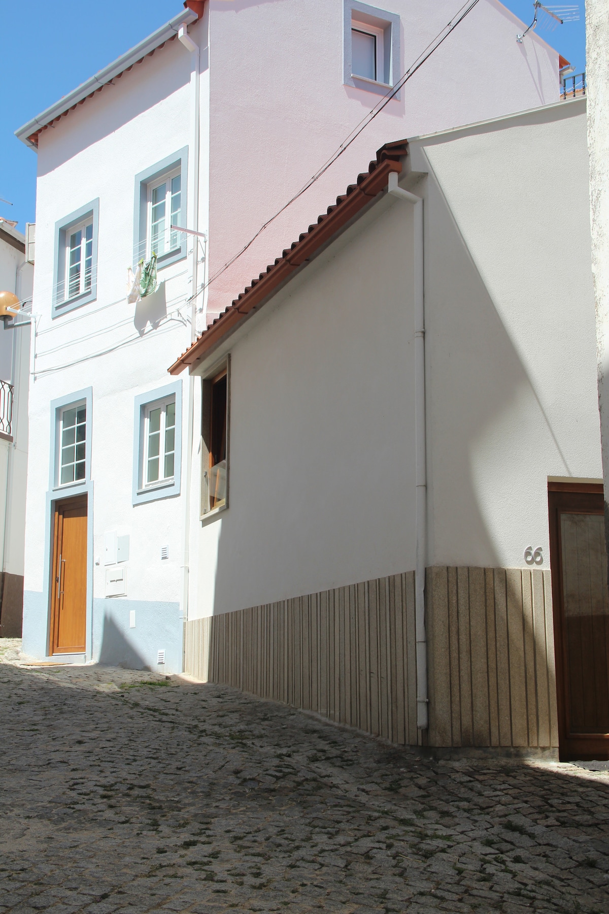 A narrow cobblestone street is flanked by charming buildings in soft pastel hues. The exterior features a mix of white and light pink walls, accented by blue window frames and wooden doors, creating a quaint and inviting atmosphere in the historic village.