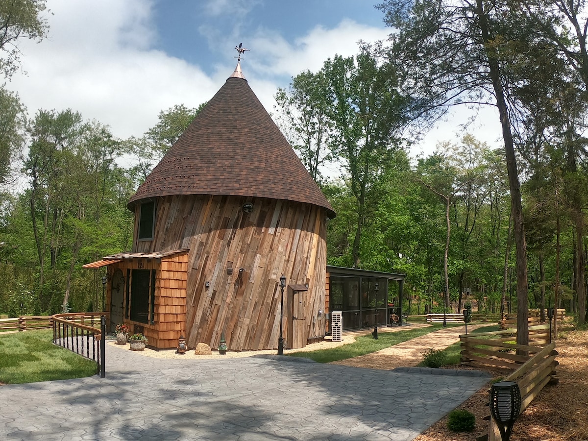 The exterior of a uniquely shaped round house, constructed with natural wood and featuring a conical roof, is surrounded by lush greenery. A spacious stone pathway leads up to the entrance, complemented by landscaped areas with various plants. Sunlight filters through the trees, enhancing the serene setting.