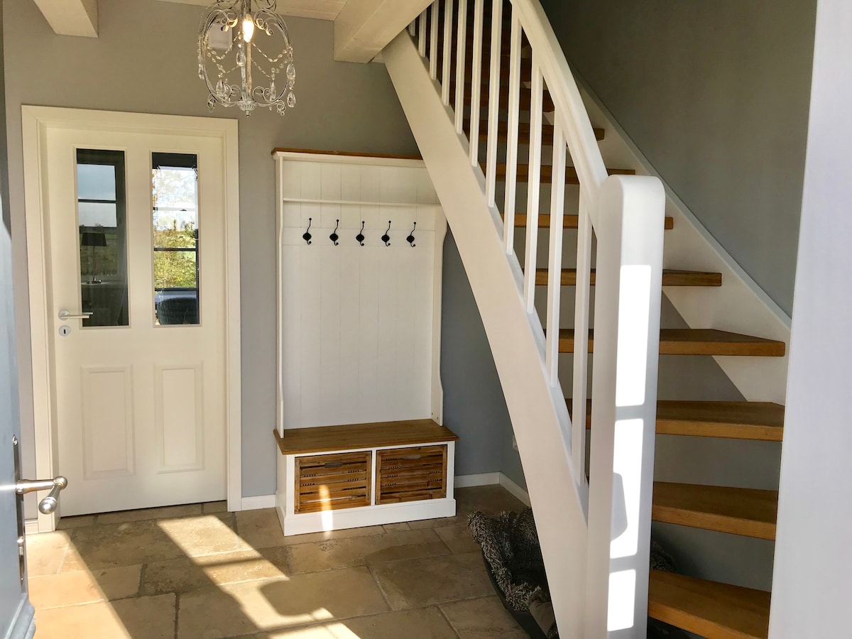 A well-lit entrance features a sliding door with glass panels and a staircase with wooden steps. A bench with storage is situated below a wall-mounted coat rack containing several hooks. The stone flooring adds a natural element to the space.