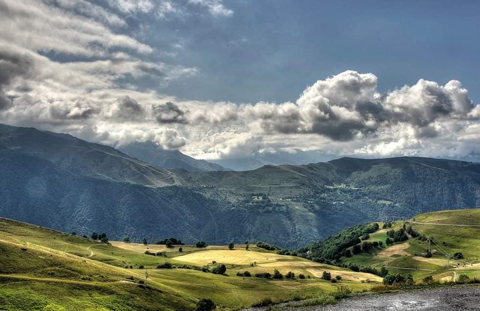 A scenic view of rolling green hills, interspersed with patches of meadows and trees. Majestic mountains tower in the background under a partly cloudy sky, creating a serene atmosphere. The terrain suggests a tranquil environment suitable for outdoor activities.