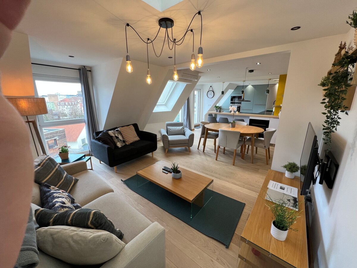 A spacious living area features a blend of comfortable seating, including a light gray sofa and a dark couch. A wooden coffee table sits centrally, complemented by plants and decor. Large windows allow natural light, illuminating the modern decor and wooden flooring.