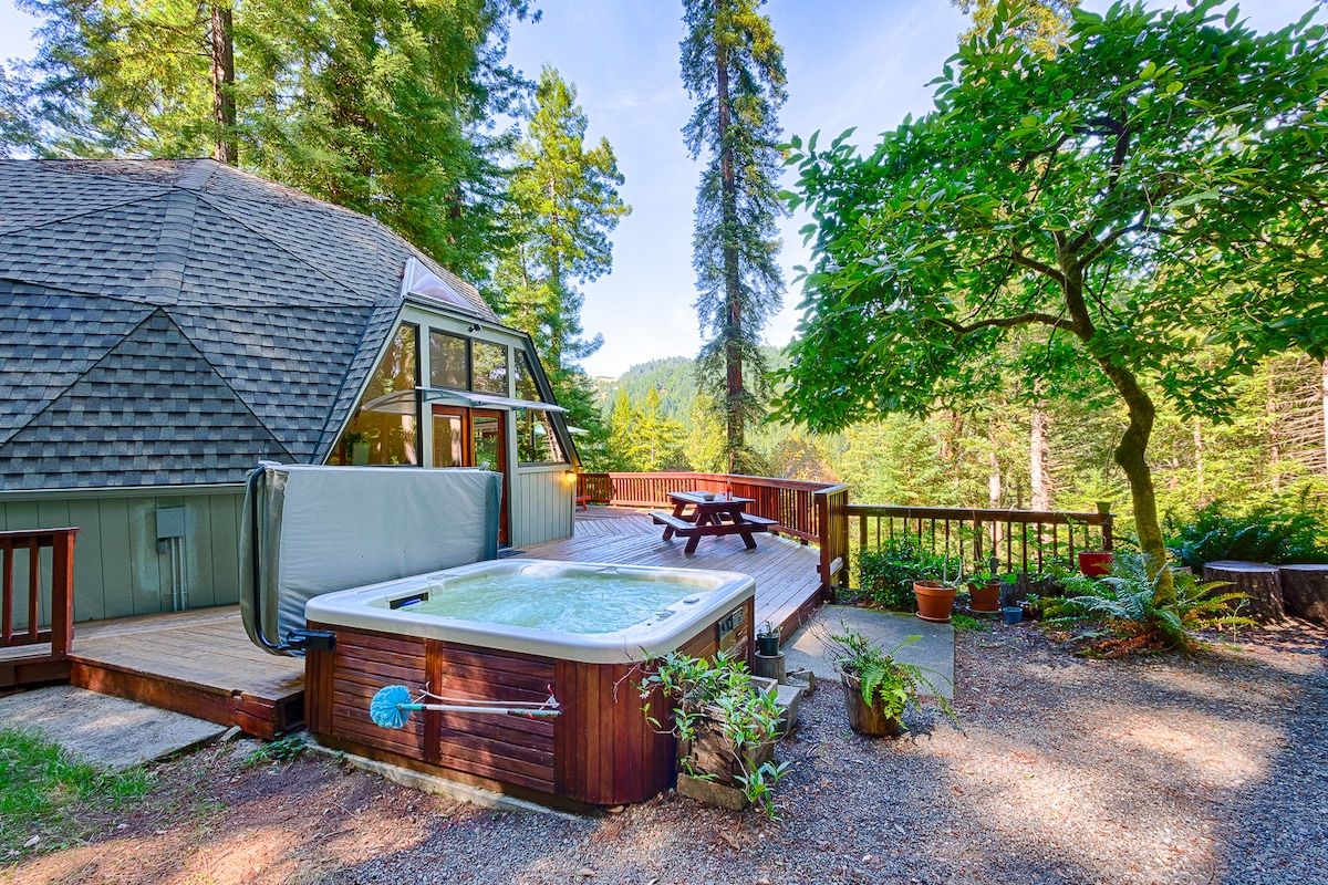 A serene outdoor space features a private hot tub on a deck surrounded by lush greenery. A picnic table is visible on the deck, inviting al fresco dining. The unique geodesic dome structure blends harmoniously with the natural landscape, providing a peaceful retreat.