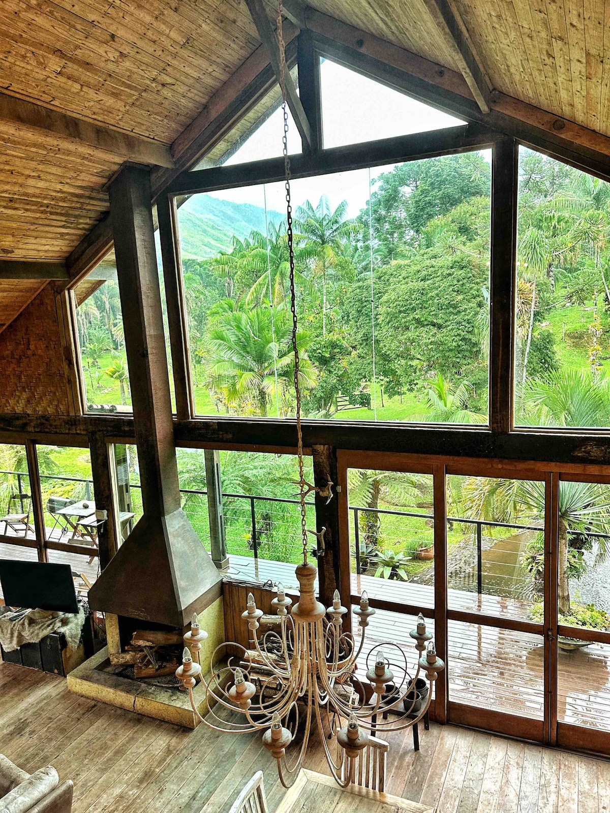 An open living space features a high wooden ceiling with a prominent chandelier hanging above. Large glass doors provide a view of the lush greenery outside, blending indoor comfort with the natural surroundings.