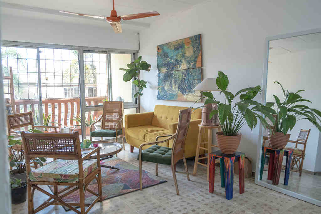 A bright living room features a yellow sofa and several chairs arranged around a glass table. Large windows allow natural light and the space is adorned with indoor plants. A colorful wall art piece and a mirror enhance the room's welcoming aesthetic.