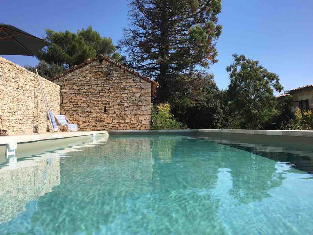 A clear swimming pool is set against a backdrop of rustic stone walls and greenery. Sunlight reflects off the water's surface, enhancing the serene environment. A lounge chair is positioned nearby, inviting relaxation. Towering trees provide shade in the surrounding area.