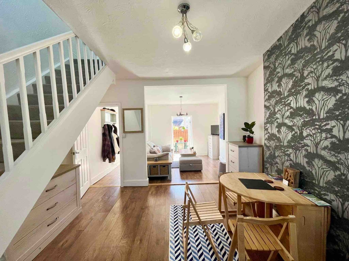A light-filled area showcases a cozy dining space with wooden furniture, set against a patterned wallpaper. A staircase is visible on the left, leading to upper levels. The open layout invites views into a living area with comfortable seating and access to the backyard.