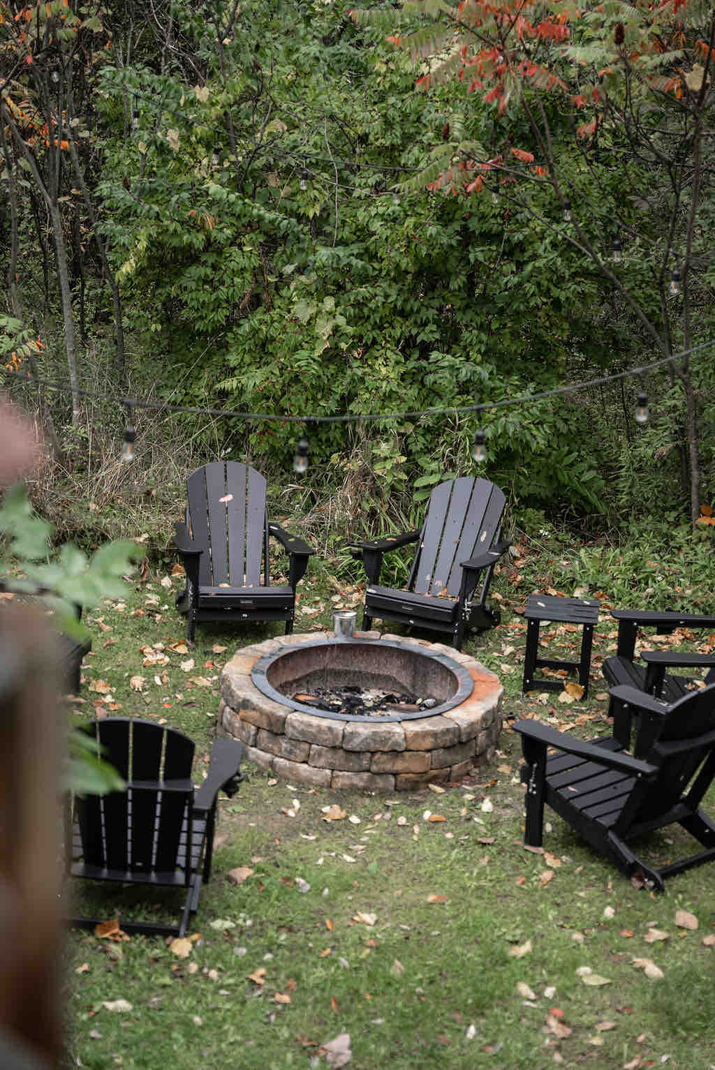 A cozy fire pit area is surrounded by five black Adirondack chairs and a small side table. The stone fire pit is centered on a grassy patch, with trees and autumn foliage providing a natural backdrop. Soft lighting is visible above, enhancing the outdoor setting.