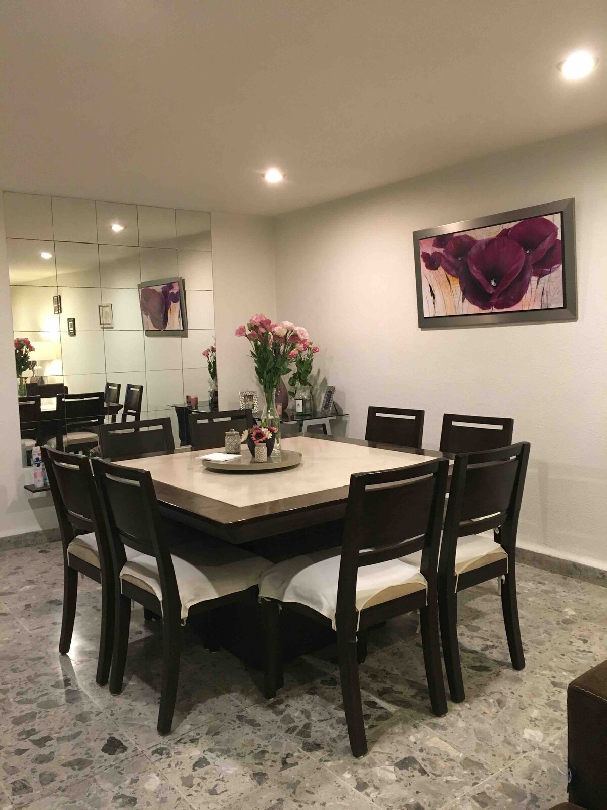 A dining area is presented with a rectangular table surrounded by six dark wooden chairs, each featuring soft seat cushions. A decorative arrangement of flowers is placed in the center. A mirror reflects the space, enhancing the openness, while a framed artwork adorns the wall.