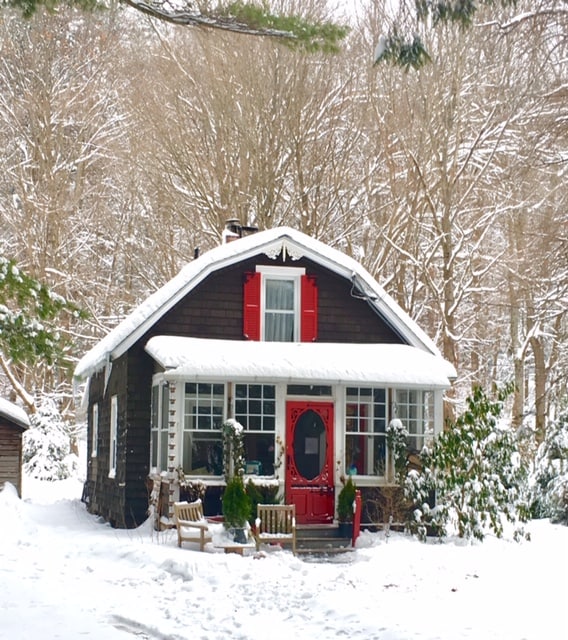 A cozy cottage exterior is framed by a snowy landscape. Red shutters and a red door stand out against the dark wood facade. Snow blankets the ground, and evergreen shrubs are visible, enhancing the tranquil winter scene.