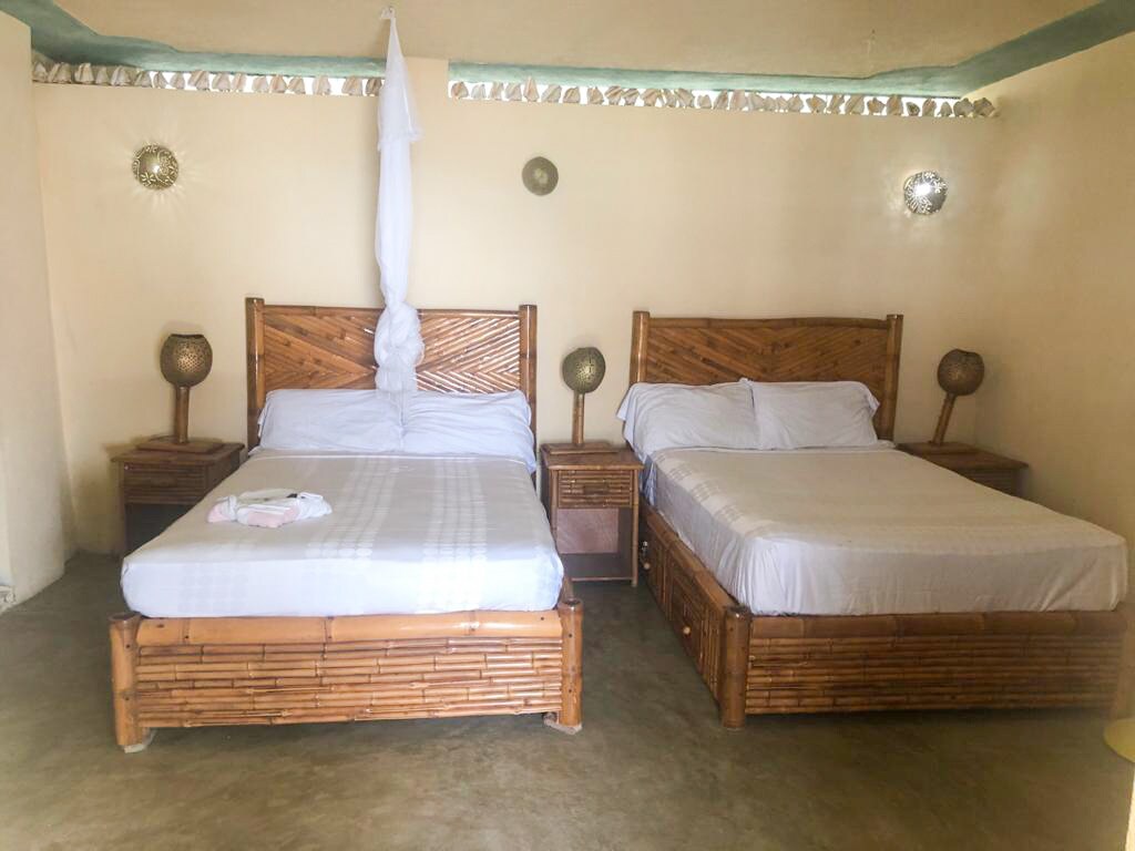 A cozy bedroom features two bamboo-style beds with light bedding, complemented by bedside tables and lamps. The walls are painted in a soft hue, contributing to a calming environment. Natural light gently enters, highlighting the simplicity of the decor.