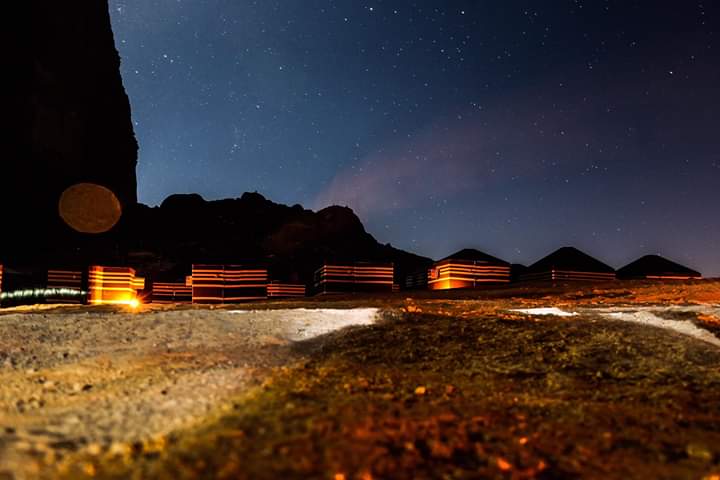 The image captures a clear night sky illuminated by stars above a campsite. Bedouin tents are arranged in rows, subtly lit by warm lights. The sandy ground reflects the glow, while the silhouette of a mountain creates a natural backdrop, enhancing the serene atmosphere of the setting.