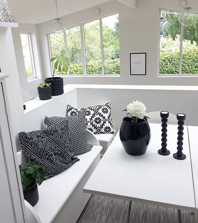 A bright and airy dining area is showcased with large windows allowing natural light to enter. A minimalist white table is accompanied by an upholstered bench and decorative pillows in black and white patterns. A vase with a flower and candles serves as the centerpiece.