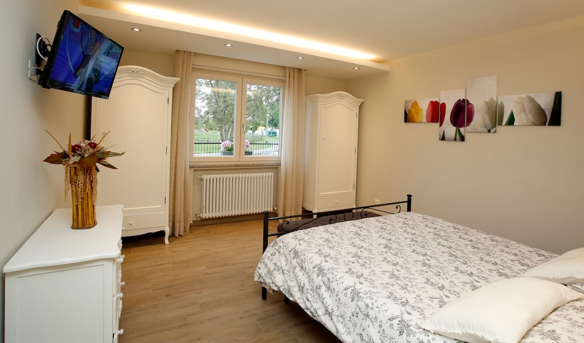 A spacious bedroom features a dark metal bed with a floral-patterned comforter. Two white wardrobes are positioned beside the bed, and a television is mounted on the wall. Natural light enters through a large window, framed by soft curtains, overlooking a green area.
