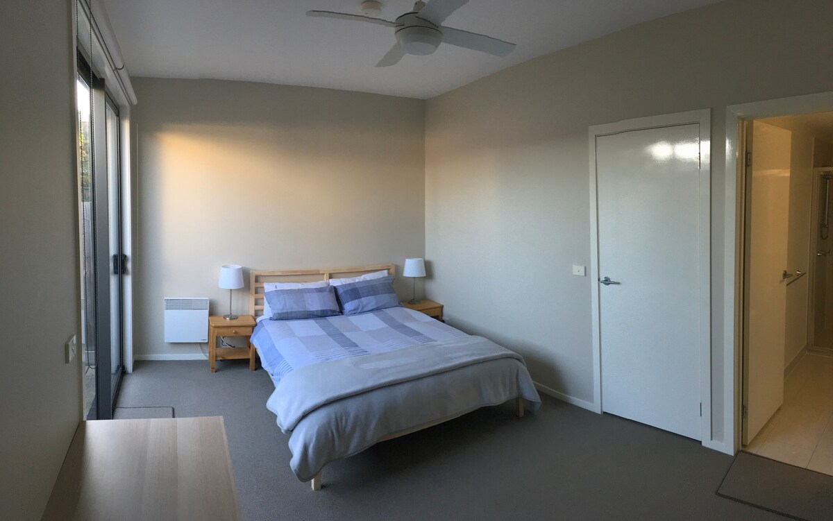 A clean and tidy private room features a spacious bed flanked by two bedside tables with lamps. A ceiling fan is mounted above, and natural light filters through large windows leading to an outdoor courtyard. A door provides access to an ensuite bathroom.