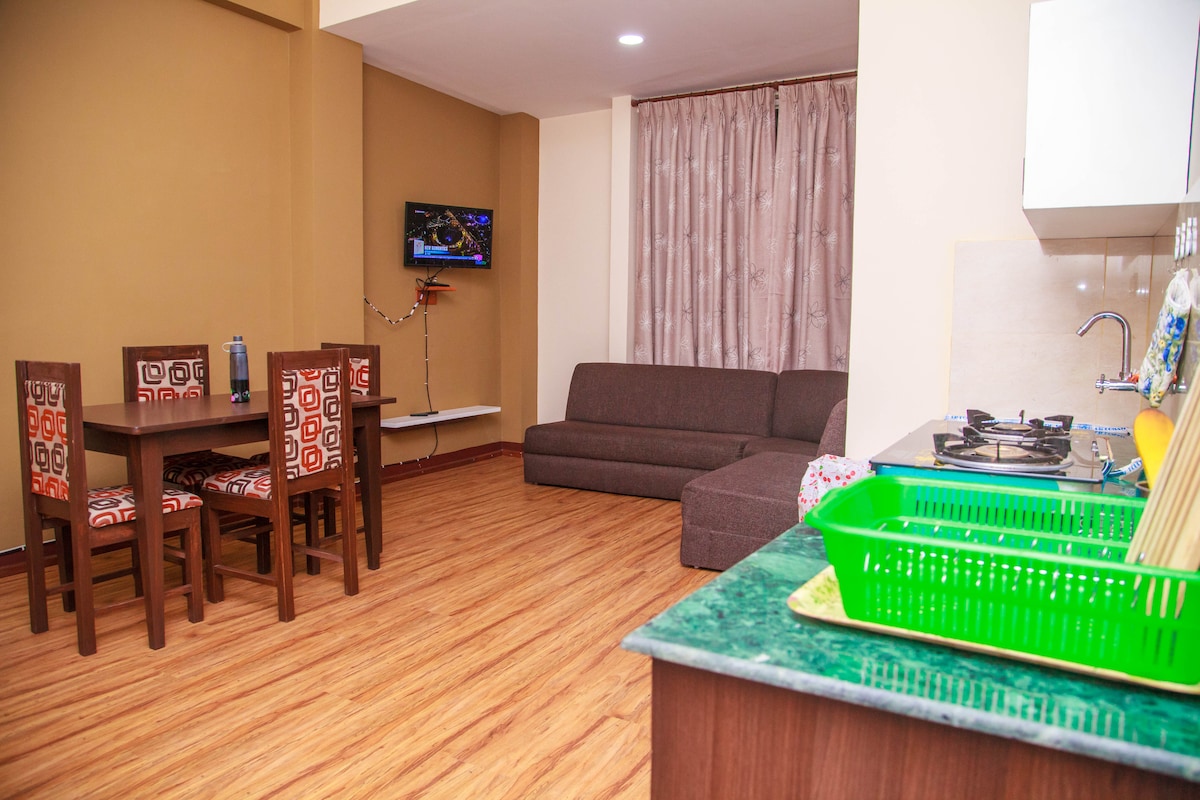 A functional living space is displayed, featuring a dining table with four chairs, a comfortable sofa, and a wall-mounted television. The kitchenette includes a green drying rack and is complemented by a countertop in a striking green hue. Soft lighting enhances the warm atmosphere.